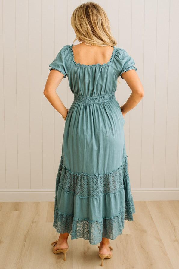 Woman wearing a teal dress with lace details, standing against a white paneled wall.
