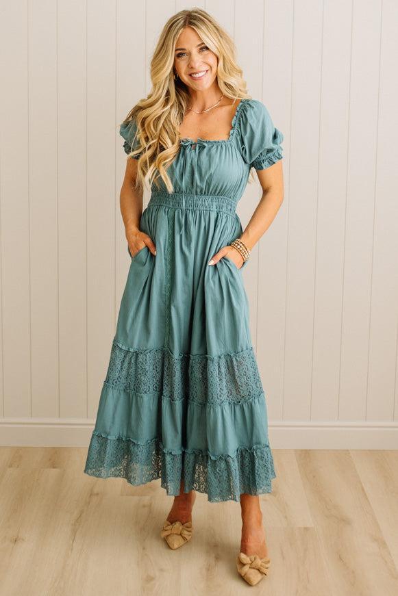 Woman wearing a teal dress with lace details against a light wooden paneled wall.