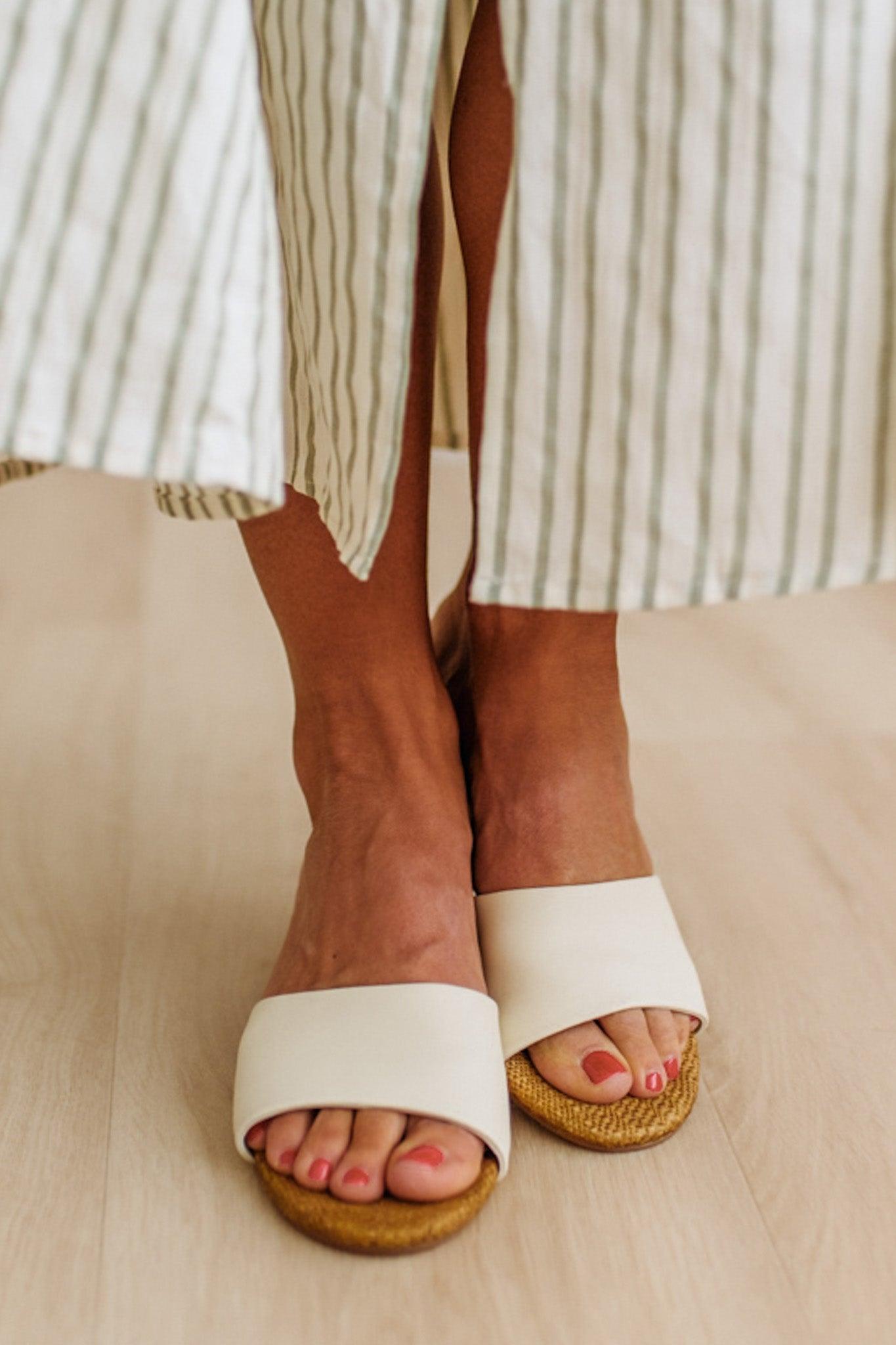 White sandals with raffia soles worn by a person sitting on a wooden floor.