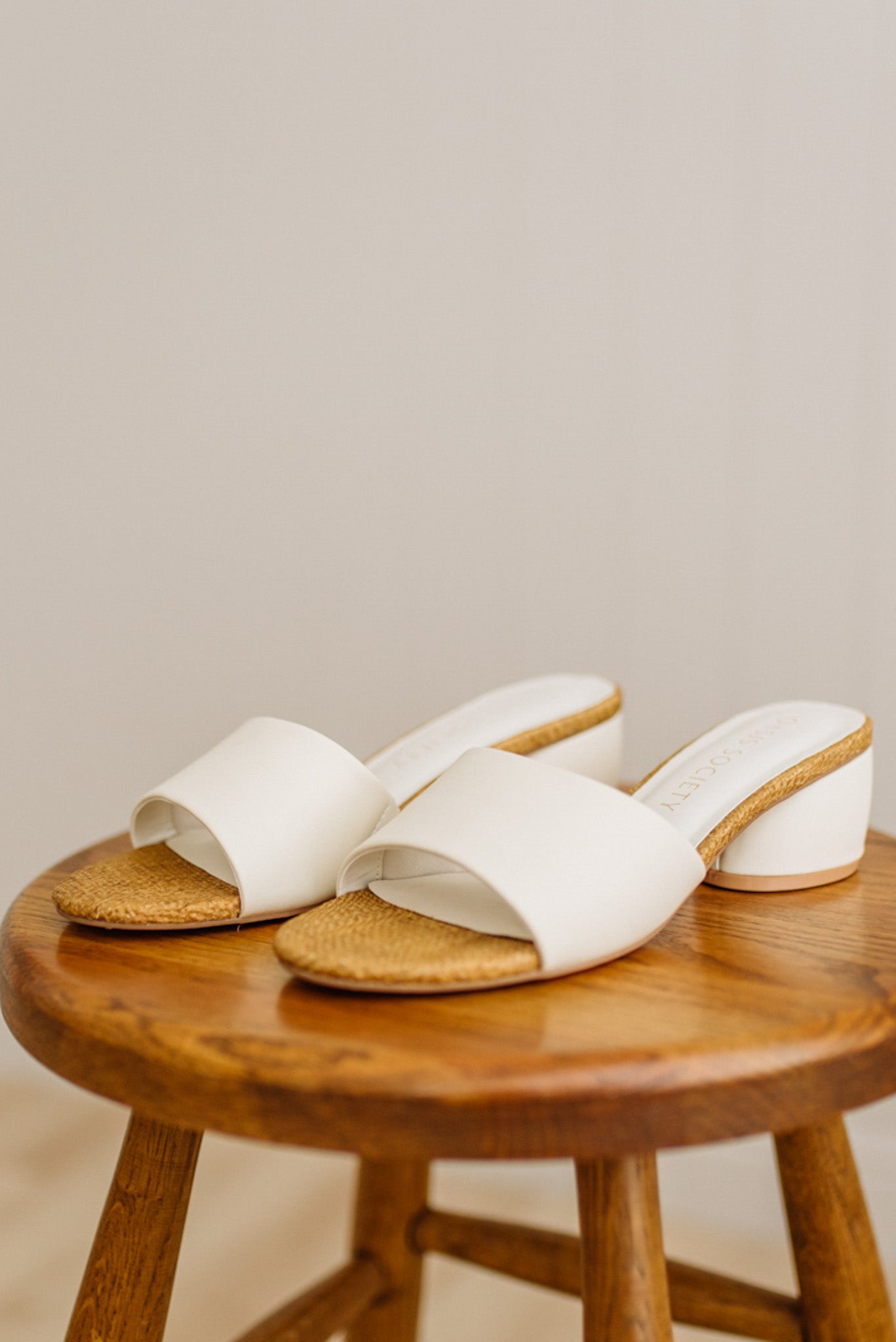 White sandals with raffia soles on a wooden stool against a neutral background