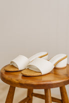 White sandals with raffia soles on a wooden stool against a neutral background