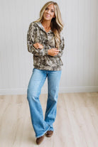 A woman stands indoors wearing a camouflage-patterned hoodie and light blue jeans, with her arms crossed. The background shows a white paneled wall and light wood flooring.