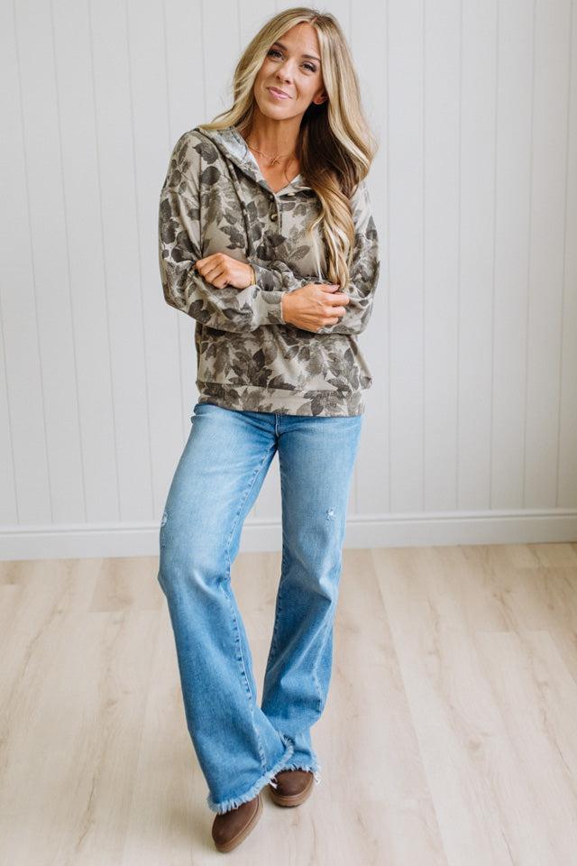A woman stands indoors wearing a camouflage-patterned hoodie and light blue jeans, with her arms crossed. The background shows a white paneled wall and light wood flooring.