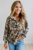 A woman wearing a long-sleeve pullover hoodie with a brown and beige leaf pattern, paired with blue jeans, standing against a white paneled wall.