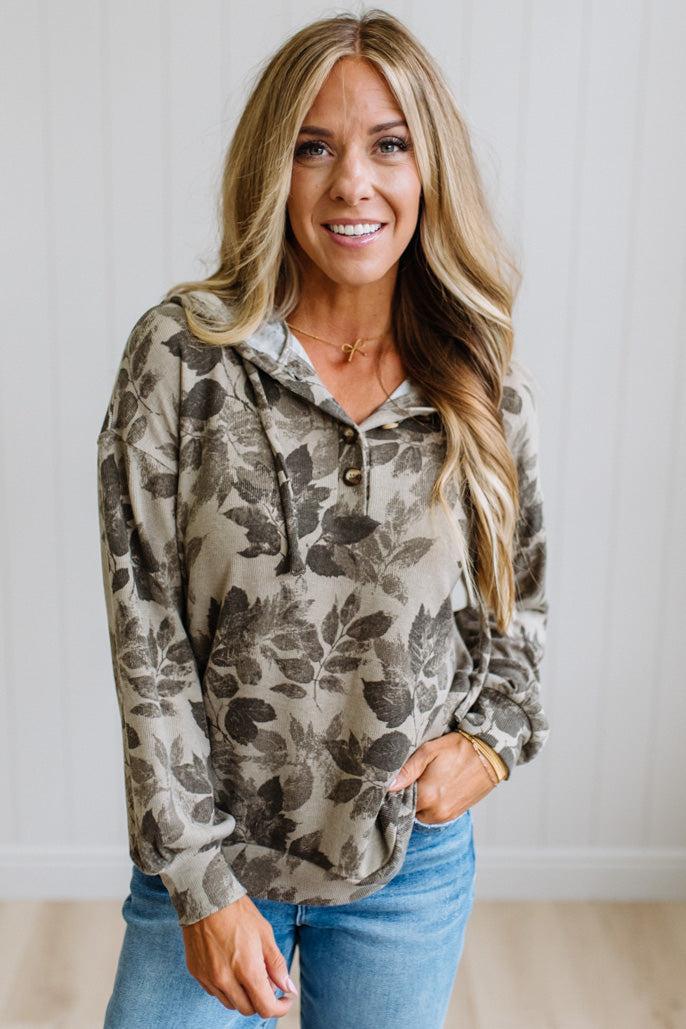 A woman wearing a long-sleeve pullover hoodie with a brown and gray floral pattern on a beige background, paired with blue jeans, standing against a white paneled wall.