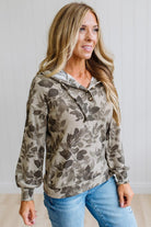 A woman wearing a beige and brown leaf-patterned pullover hoodie with blue jeans, standing against a white paneled wall.
