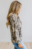A woman wearing a long-sleeve hooded sweatshirt with a distressed, faded floral pattern in beige and brown, paired with blue jeans, standing against a white paneled wall.
