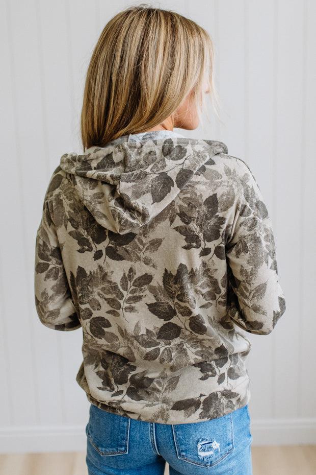 A woman viewed from behind wearing a beige hooded sweatshirt with a dark brown leaf pattern, paired with blue jeans, set against a white paneled wall.