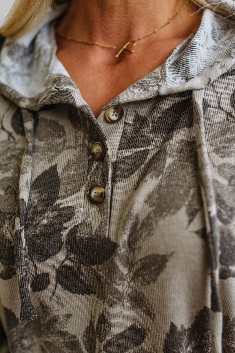 Close-up of a person wearing a hooded pullover top with a muted gray and beige leaf print, featuring a button placket and drawstring hood.