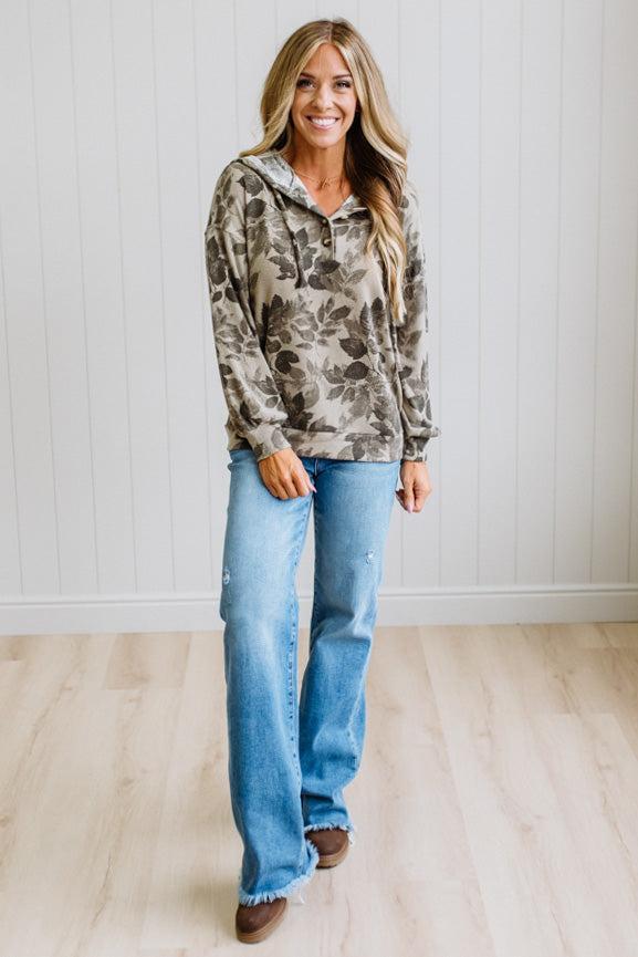 A woman modeling a long-sleeve hooded pullover with a brown and beige floral pattern, paired with light-wash blue jeans and brown shoes, standing in a bright room with a white paneled wall and light wood floor.