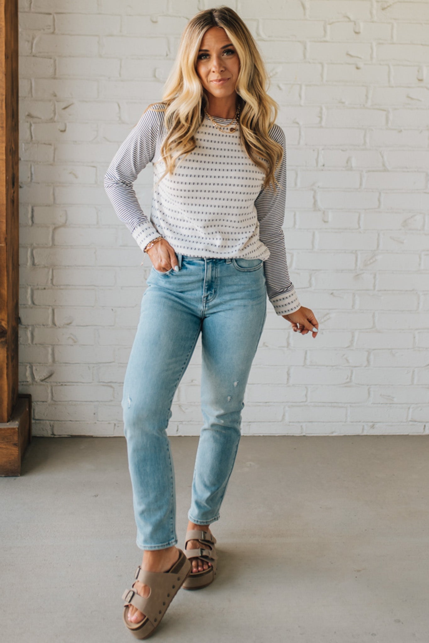 Woman wearing a patterned top and light blue jeans against a white brick wall.