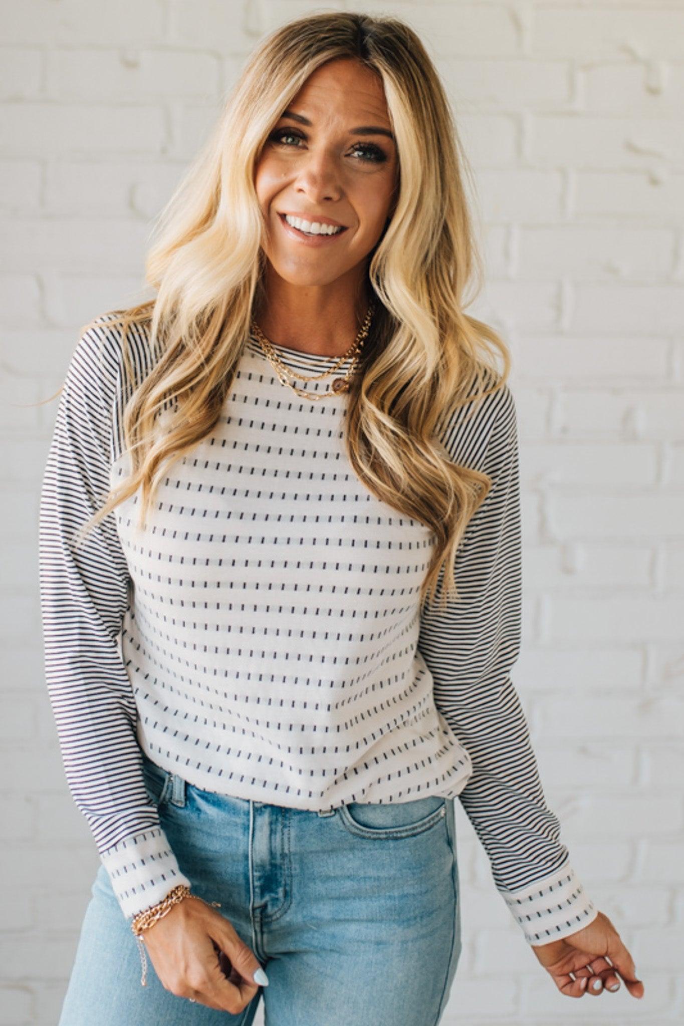 Woman wearing a striped long-sleeve top and jeans against a white brick wall.
