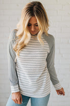 Woman wearing a long-sleeve patterned top against a white brick wall.