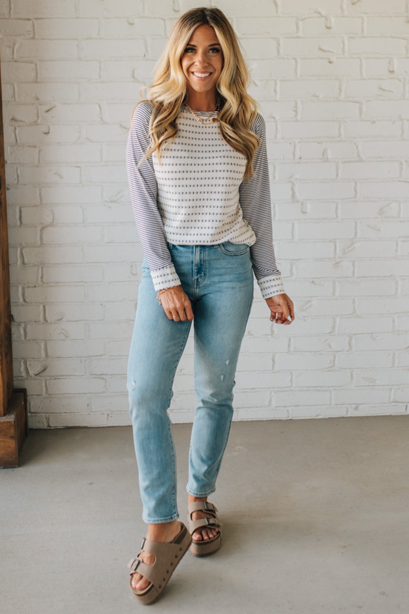 Woman wearing a striped shirt and jeans standing against a white brick wall.