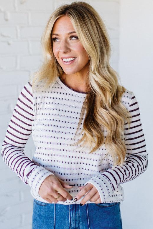 Woman wearing a striped sweater and blue jeans against a white brick wall.