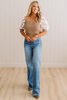 A woman modeling a beige ribbed knit top with white lace puff sleeves and blue flare jeans, standing in a room with light wood floors and white paneled walls.