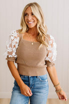 A smiling blonde woman wearing a beige ribbed knit top with white floral lace puff sleeves, paired with blue jeans, standing against a light neutral paneled wall.
