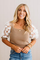A smiling blonde woman wearing a beige ribbed knit top with white floral lace puff sleeves, paired with blue jeans, against a light neutral background.