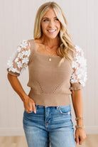 A smiling blonde woman wearing a beige ribbed knit top with white floral lace puff sleeves, paired with blue jeans, standing against a light beige paneled wall.