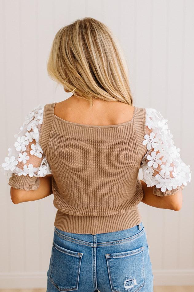 A woman viewed from behind wearing a tan ribbed knit top with white floral lace puff sleeves, paired with blue jeans, against a light beige paneled wall.