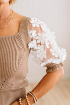 A close-up of a woman wearing a taupe ribbed knit top with short puff sleeves featuring white floral lace applique.