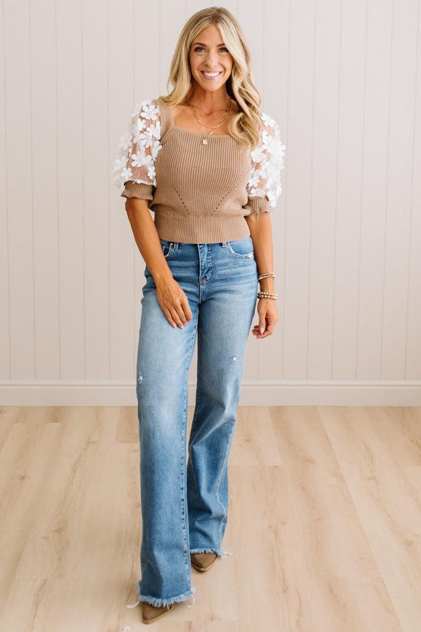 A woman modeling a beige ribbed knit top with white lace puff sleeves and light-wash blue flare jeans, standing in a room with light wood floors and white paneled walls.