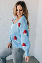 Light blue knit cardigan with charming red and white mushroom appliques on the sleeves and body, on a blonde woman.