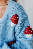 Light blue knit cardigan with charming red and white mushroom appliques on the sleeves and body, on a blonde woman.