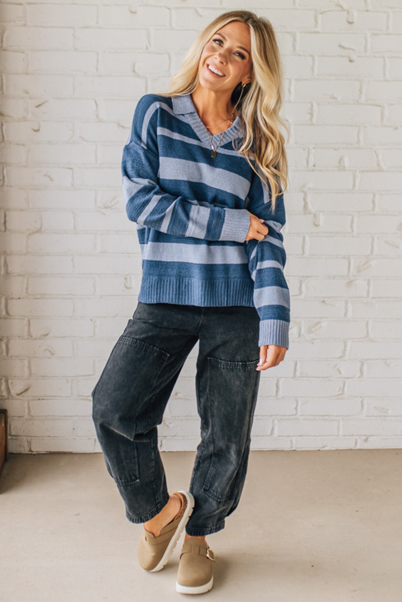 Woman wearing a blue striped sweater and dark pants against a white brick wall.