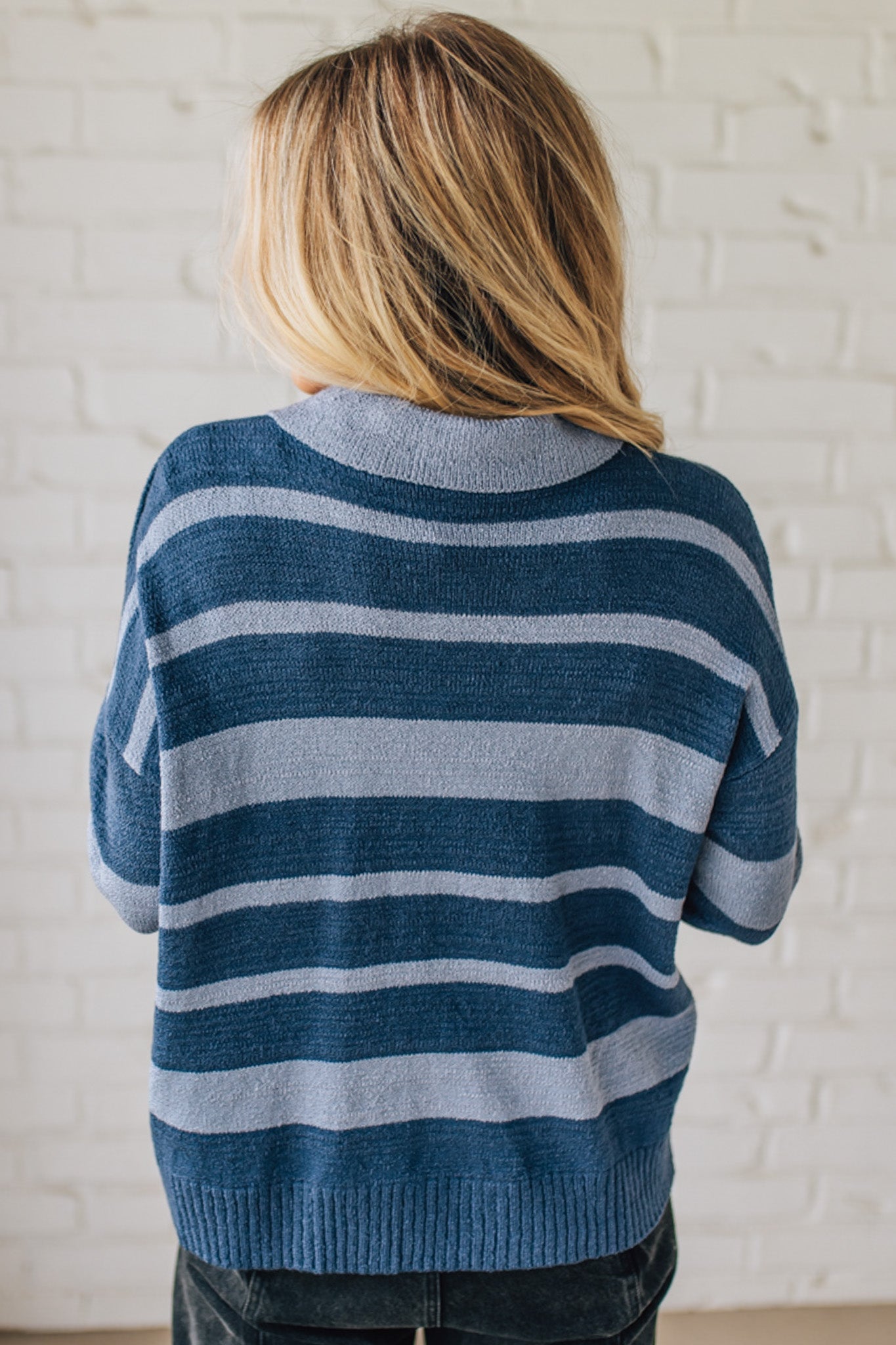 Person wearing a blue and light blue striped sweater against a white brick wall.