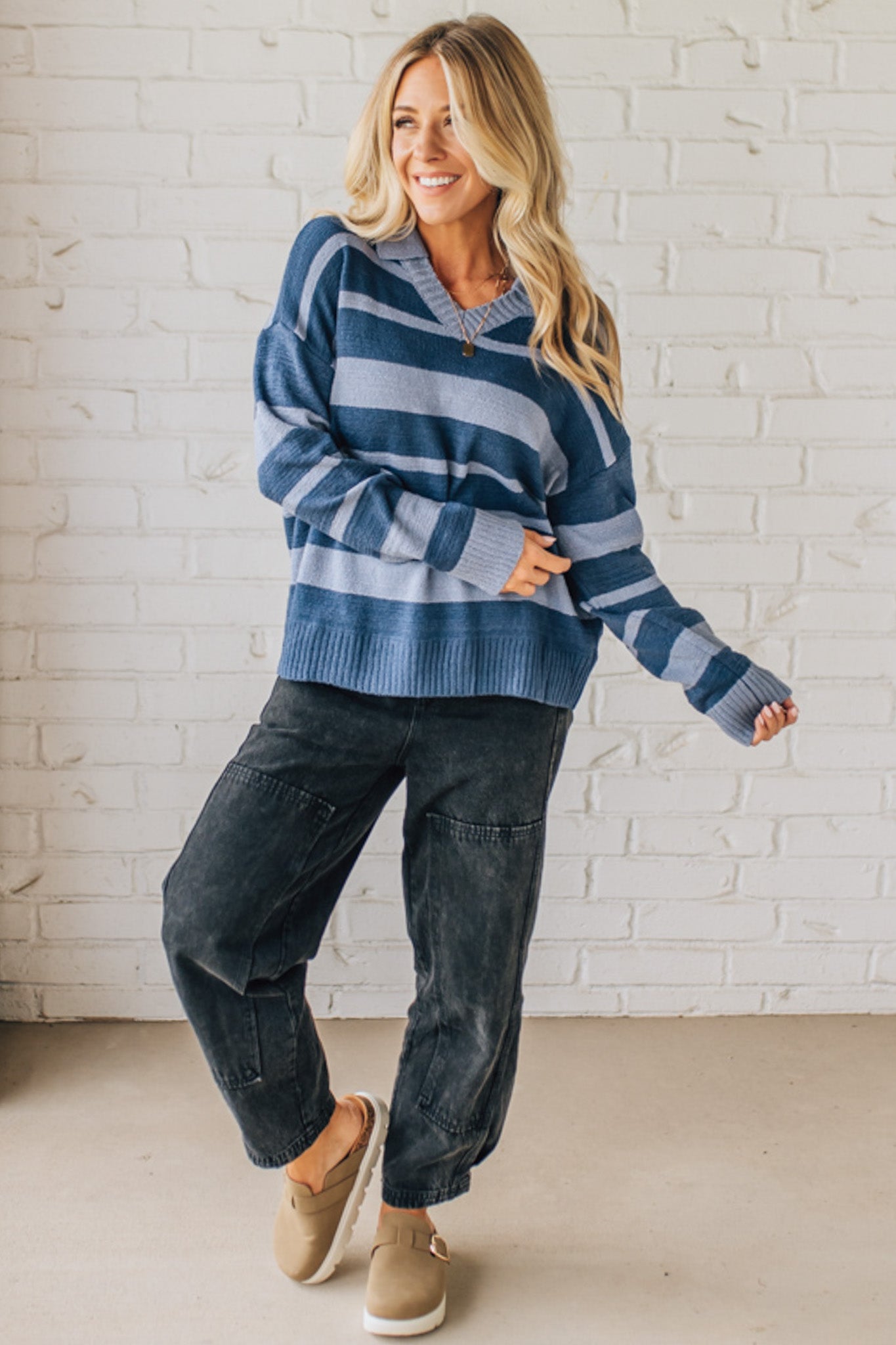 Woman wearing a blue striped sweater and dark pants against a white brick wall.