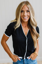 A smiling blonde woman wearing a black short-sleeve button-front cardigan with white trim, paired with blue jeans, standing against a light neutral wall.