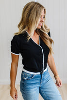 A woman wearing a black short-sleeve button-front top with white trim and light blue jeans, standing against a light indoor wall.