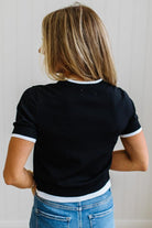 A woman viewed from behind wearing a black short-sleeve ringer t-shirt with white trim on the collar and sleeve cuffs, paired with blue jeans.