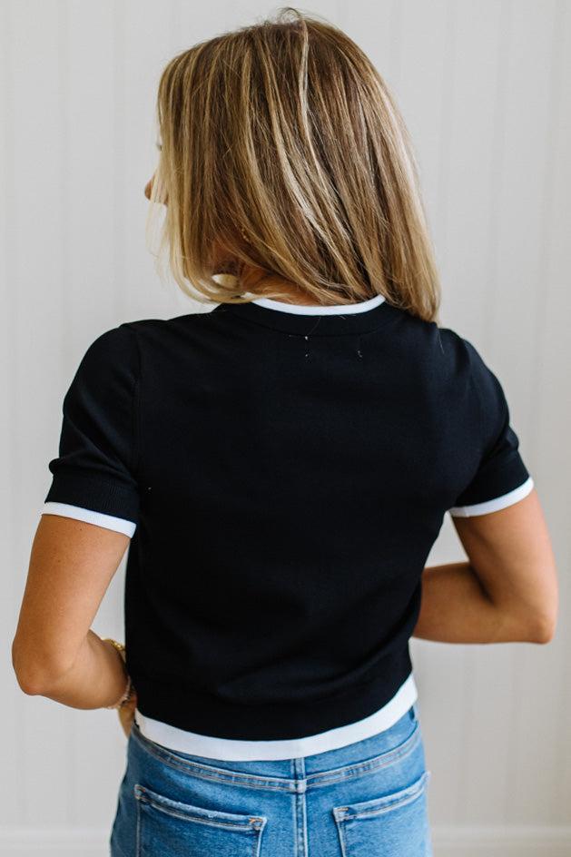 A woman viewed from behind wearing a black short-sleeve ringer t-shirt with white trim on the collar and sleeve cuffs, paired with blue jeans.