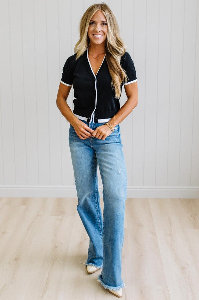 A woman modeling a black short-sleeve knit top with white trim and light-wash blue flare jeans, standing in a bright room with a white paneled wall and light wood floor.