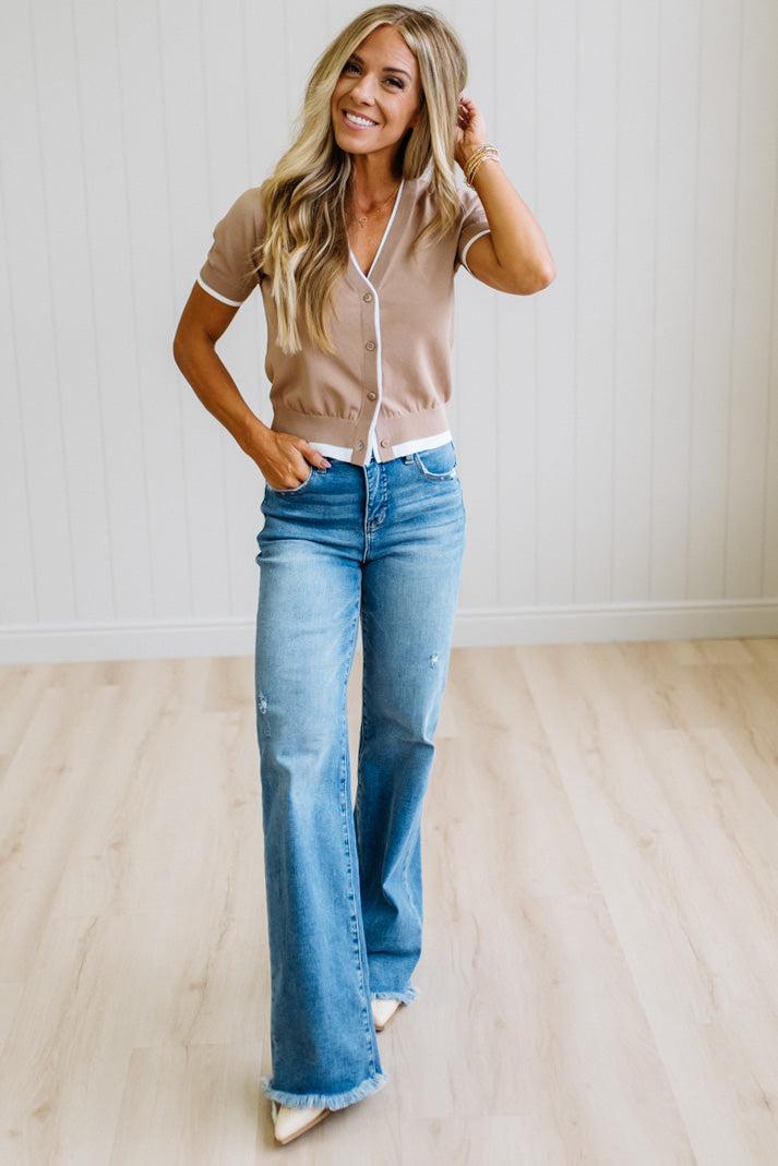 A woman modeling a beige short-sleeve button-front cardigan with white trim over a white top, paired with blue flare jeans and beige pointed shoes, standing in a bright room with light wood floors and white paneled walls.