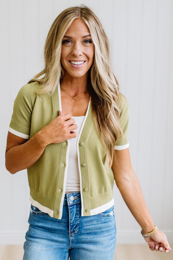 A smiling blonde woman wearing a short-sleeve olive green knit cardigan with white trim over a white tank top and blue jeans, standing against a light paneled wall.