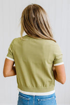 A woman viewed from behind wearing a cropped sage green short-sleeve top with white ribbed trim at the collar, cuffs, and hem, paired with blue jeans against a white paneled wall.