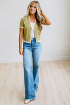 A woman modeling a light olive green short-sleeve cropped cardigan with white trim, worn over a white top and blue wide-leg jeans, standing in a room with light wood floors and white paneled walls.