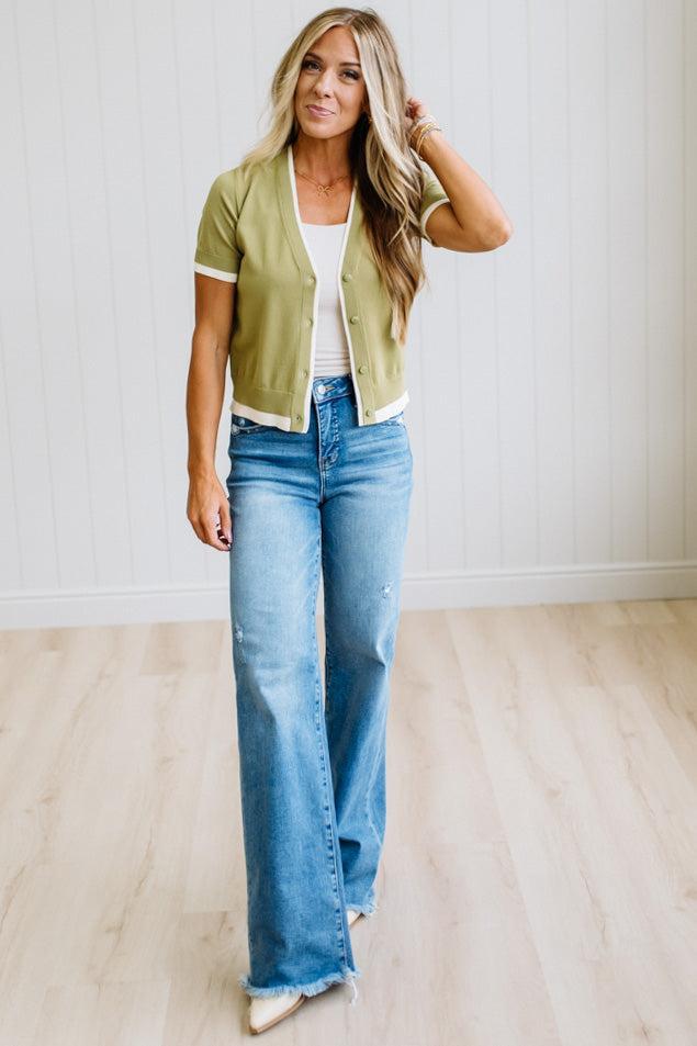 A woman modeling a light olive green short-sleeve cropped cardigan with white trim, worn over a white top and blue wide-leg jeans, standing in a room with light wood floors and white paneled walls.