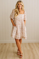 Woman wearing a floral dress standing against a light wooden paneled wall.
