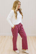 A woman modeling a pair of burgundy and white polka-dot lounge pants with a white long-sleeve top, standing in a room with light wood floors and white paneled walls.