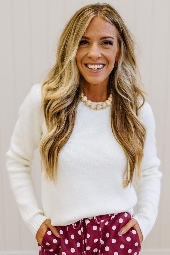 A smiling woman with long blonde wavy hair wearing a white knit sweater, a chunky pearl necklace, and burgundy pants with white polka dots, standing against a light beige paneled wall.