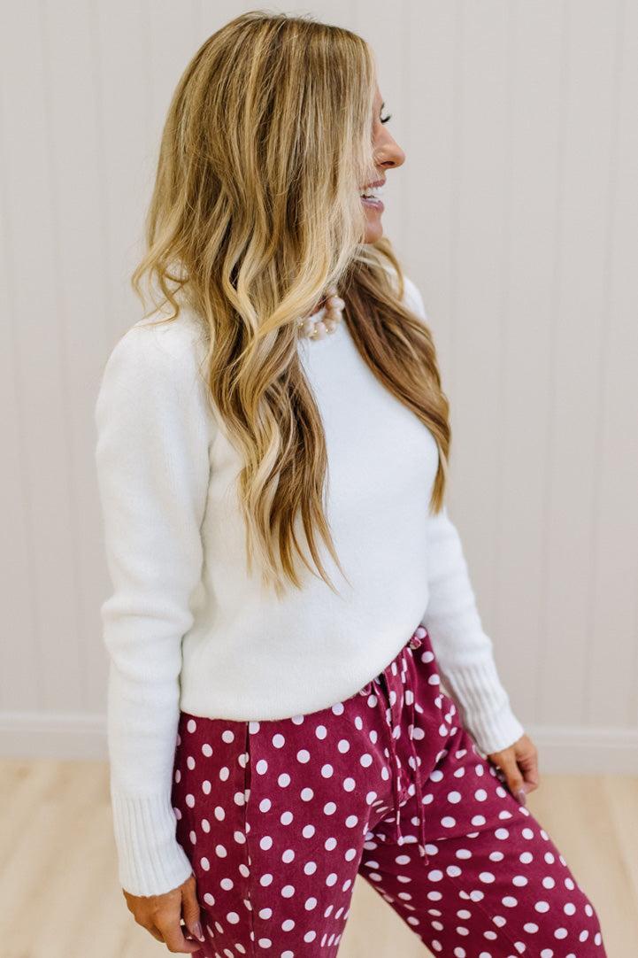 A woman wearing a white long-sleeve knit sweater and burgundy pants with white polka dots, shown from the side in a brightly lit indoor setting.