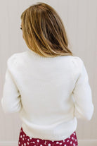 A woman viewed from behind wearing a white knit sweater and burgundy pants with white polka dots, set against a light beige paneled wall.