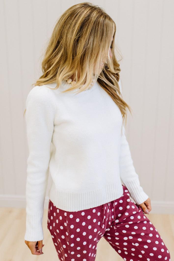 A woman wearing a white long-sleeve mock neck knit sweater and burgundy pants with white polka dots, shown from a side-back angle against a light wood-paneled wall.