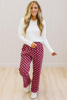 A woman modeling burgundy pants with white polka dots, paired with a white knit sweater and white shoes, standing in a room with light wood floors and white paneled walls.