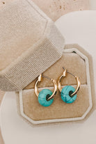 Gold hoop earrings with round turquoise stone pendants, displayed in a beige jewelry box.