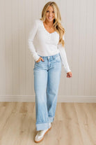 Woman wearing a white long-sleeve top and light blue jeans standing against a beige wall.